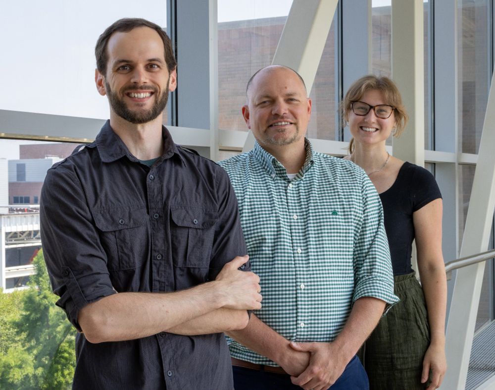 Drs Troy Burtchett, Neal Hammer, and doctoral student Jessica Lysne stand next to each other. They are in front of a window and are all smiling at the camera. 