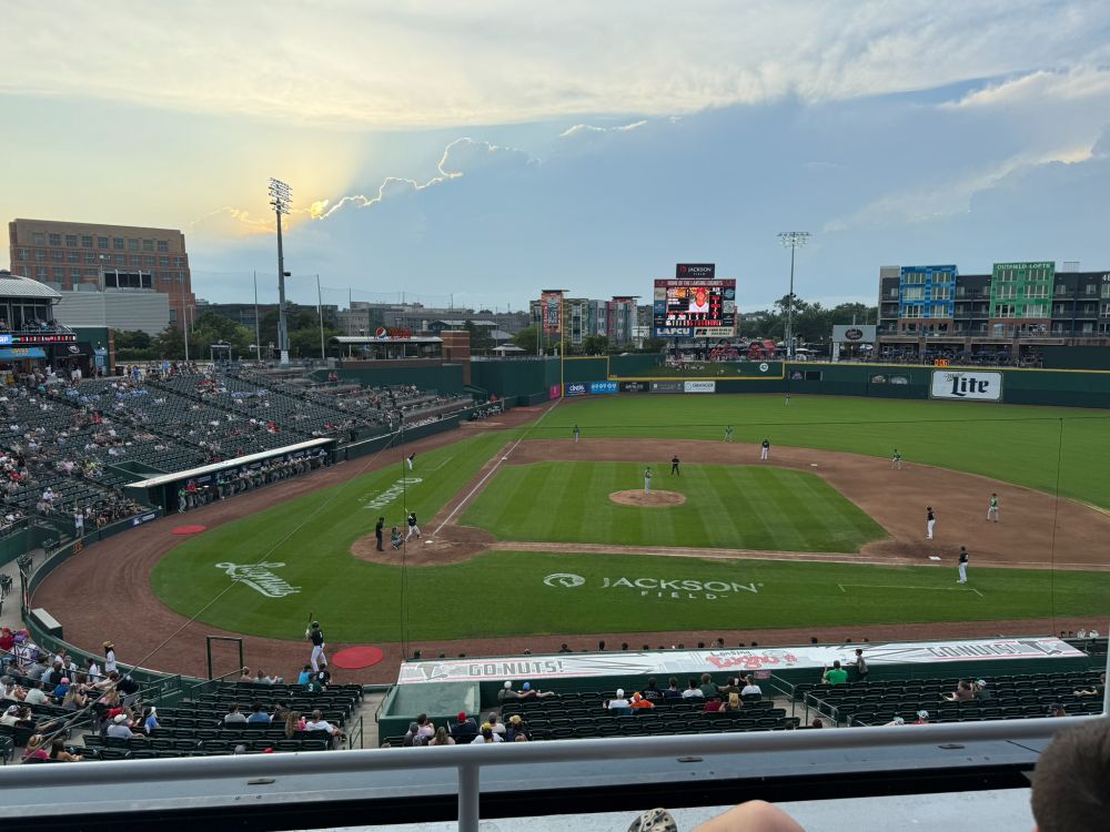 This image shows the ballfield at the Lugnuts stadium. The sun is peeking out behind some clouds and illuminating the edge of the cloudline. The teams are playing baseball on the diamond, and the 
