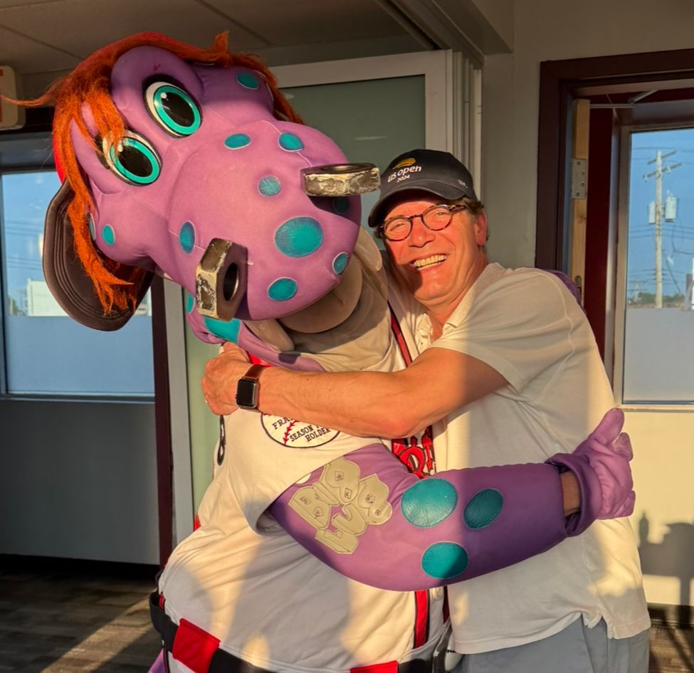 Vic DiRita hugs Big Lug, the Lugnuts mascot. Big Lug is a purple dinosaur with blue spots and lugnuts for nostrils. He has stringy orange hair. 
