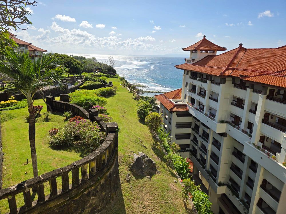 A gorgeous hotel next to the sea. It's very sunny and there is a beautiful grassy area and a palm tree. You can see the waves in the distance and it looks like cliffs down to the water.

