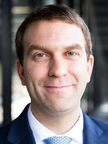 Jeff Barrick smiles at the camera. He is wearing a dark blue suit and blue patterened tie. He has short brown hair. 
