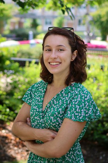 Camille Boucaud is standing in a garden and smiling at the camera. She's wearing a green and white dress and is crossing her arms. 