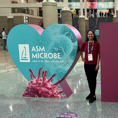 Dr. Rangarajan stands with a large, person-sized, heart-shaped sign reading "ASM Microbe." She is smiling broadly. 