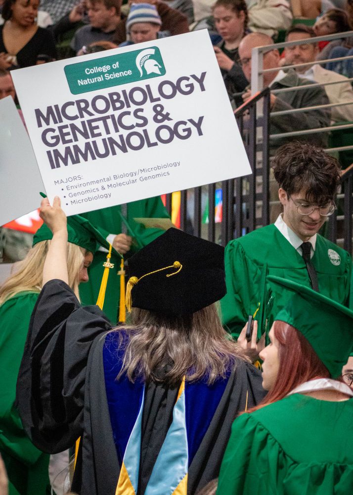 A professor in regalia with her back to the camera holds up a sign reading "Microbiology, Genetics, & Immunology."
