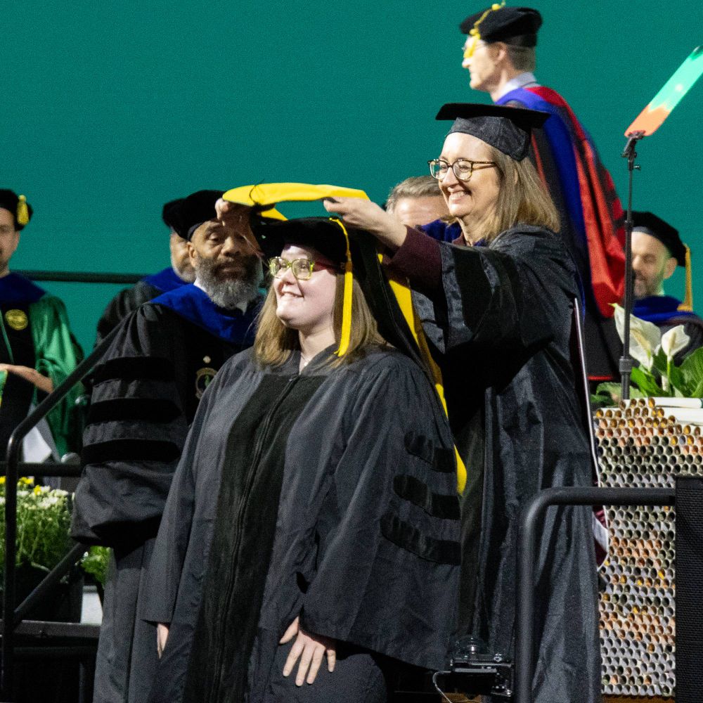 An MGI PhD student receives her hood from her advisor, Dr. Gemma Reguera. They are both smiling. 
