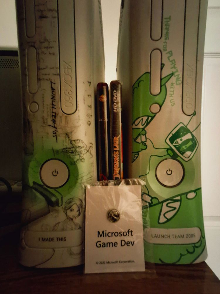 The back of the developer game pin's card which plainly reads "Microsoft Game Dev". In the background are the US Launch Team Xbox 360 which says "I made this" and a second Xbox 360 with the European Launch Team Xbox 360 faceplate on it. Also a HD-DVD copy of Dragon's Lair and the movie "300" between the Xboxes, representing what might be the full game library for HD-DVD if you count the demo on 300.