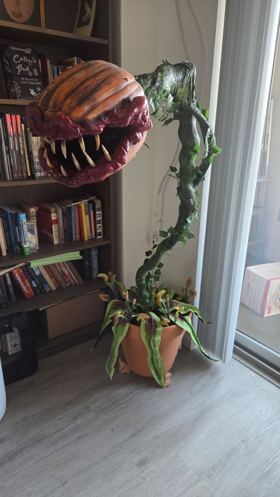 A man eating plant prop. Its base is a giant flower pot and there are big leaves sprouting from the base.  Tiny venus flytraps sprout up among the larger leaves. 