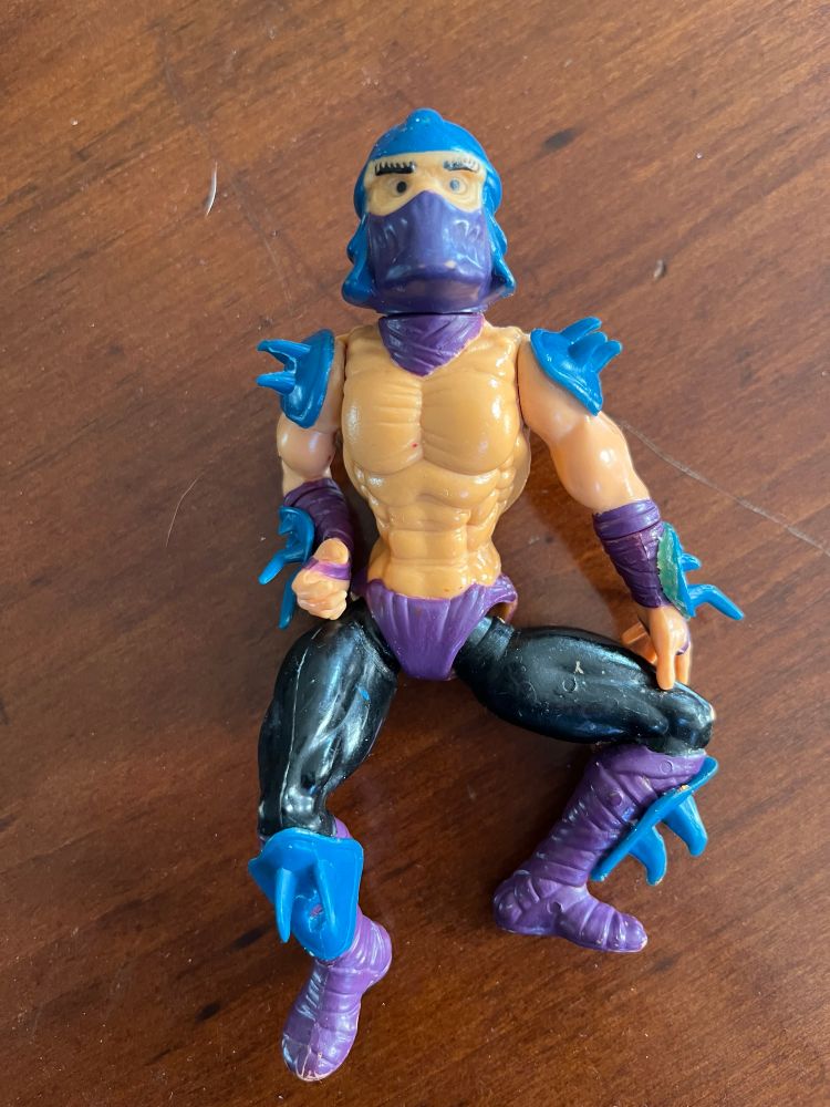 1990s Teenage Mutant Ninja Turtle action figure of Shredder. He's got blue spikes on his arms, wrists and ankles. Purple boots. Black pants. He is ripped.