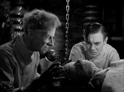 Dr Pretorius and Victo Frankenstein standing over a bandaged figure in a laboratory 