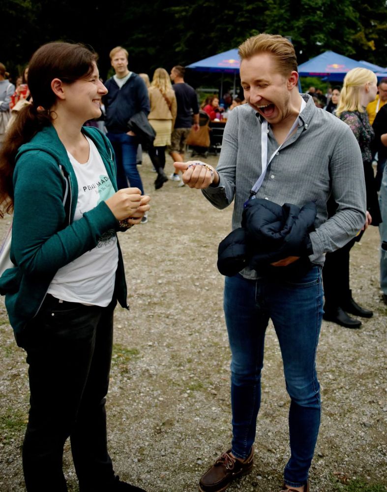 Ich stehe Sebastian (männlich, Ende zwanzig, helle Haut, blonde Haare) gegenüber. Er trägt ein graues Hemd, Jeans und einen Crew-Ausweis an einem Band um den Hals. In der rechten Hand hält er ein Armband. Sein Mund ist in einer Mischung aus Fassungslosigkeit und Begeisterung weit aufgerissen. Foto © Robert Herbe