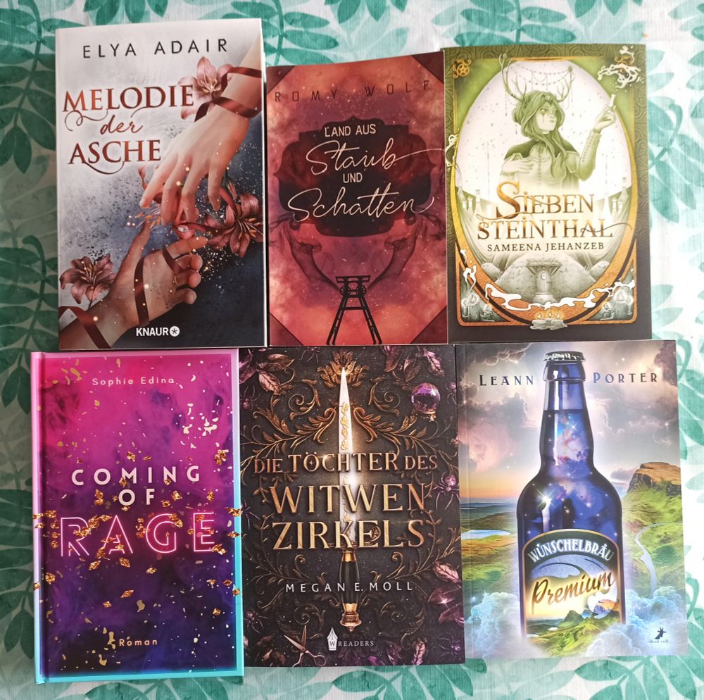 Sechs Bücher auf einer Tischdecke mit Blättermuster:
Elya Adair - "Melodie der Asche"
Romy Wolf - "Land aus Staub und Schatten"
Sameena Jehanzeb - "Siebensteinthal"
Sophie Edina - "Coming of Rage"
Megan E. Moll - "Die Töchter des Witwenzirkels"
Leann Porter - "Wünschelbräu Premium"