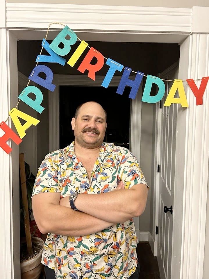 A photo of Dave on his birthday last year, wearing a colorful shirt and smiling under a sign that says "Happy Birthday"