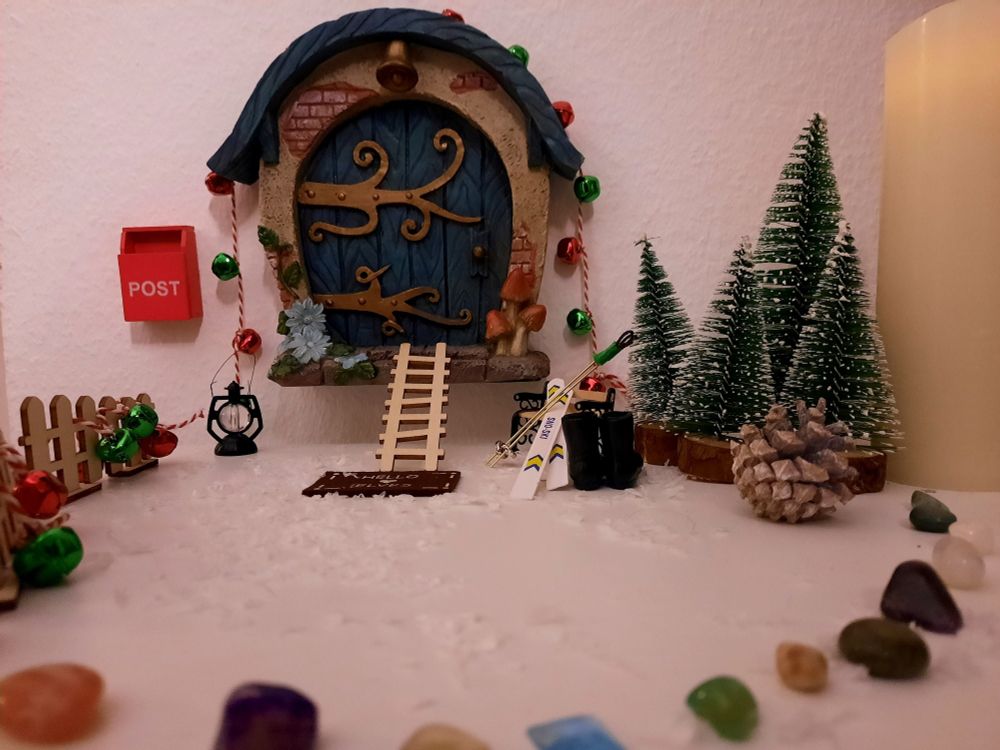 Wichteltür, davor steht jetzt eine tragbare Laterne. Es liegt Schnee und an der Bank lehnen Ski und Skistöcke. Neben der Bank stehen Stiefel.