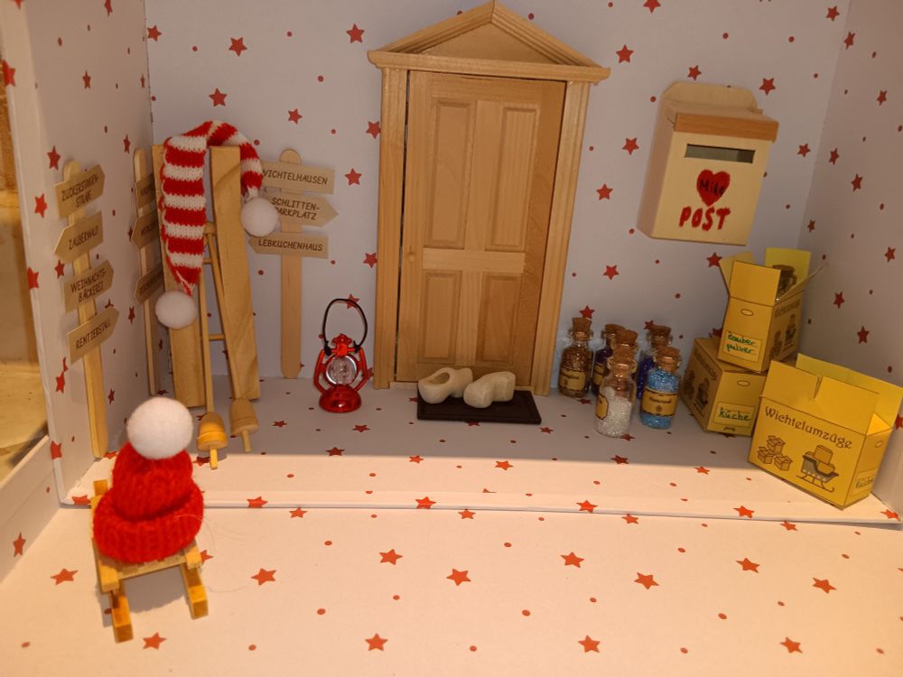 Szenerie in einem weißen Karton mit kleinen roten Sternen: mittig klebt eine Holztür, rechts daneben ein Briefkasten. Links neben der Tür befinden sich Wegweiser von Wichtelhausen bis zum Nordpol. An die Wand angelehnt sind Skier, an dem ein weiß roter Schal hängt, davor ein Schlitten mit roter Bommelmütze. Rechts von der Tür stapeln sich mehrere kleine Kisten mit der Aufschrift Wichtelumzüge. Vor der Tür liegt eine Fußmatte, darauf steht ein Paar Holzschuhe. Kleine Fläschchen mit Glitzerpulver und eine rote Laterne stehen verteilt.