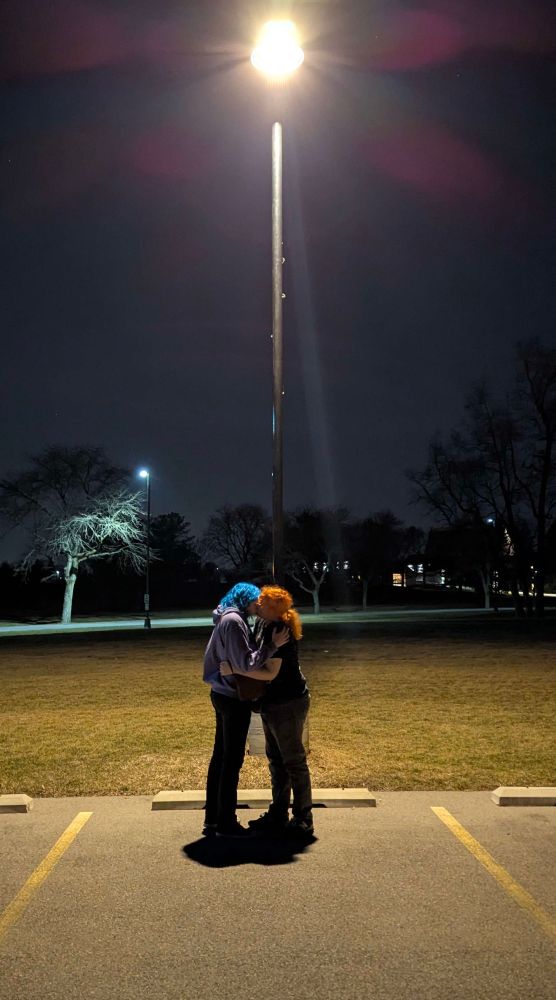 picture of two girls in a parking lot, under a streetlight. one with blue hair, one with orange. there is a bright glow above them radiating down on them, and they appear to be making out.