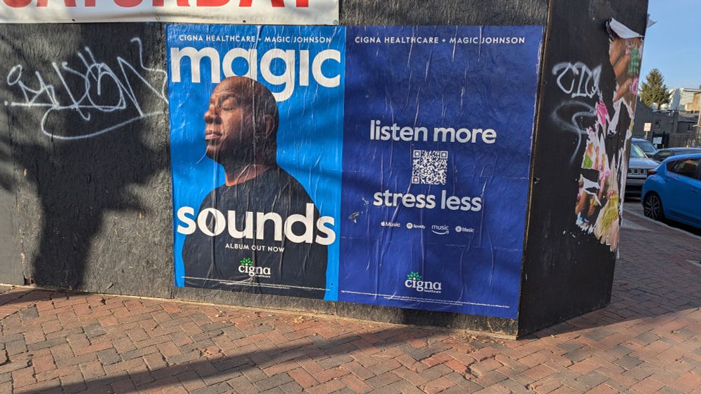 A wheat paste posted on the side of some particle board fencing at a vacant lot in the northern fells point neighborhood of Baltimore, rher advertisement is a photo of magic johnson, portrait style, with his eyes closed just kind of vibing, it is an advertisement for his lofi hip hop beats style album made with cigna healthcare called magic sounds