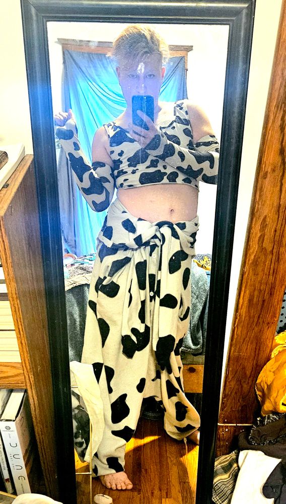 A transmasculine individual takes a mirror selfie in slightly risque cow print clothing including gloves/arm warmers. 