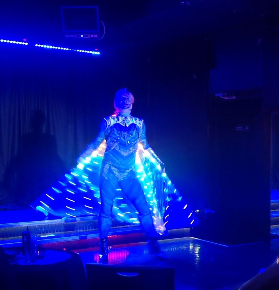 A performer stands mid stage with curtainy wings of rainbow light extended in an etherial blue wash. 