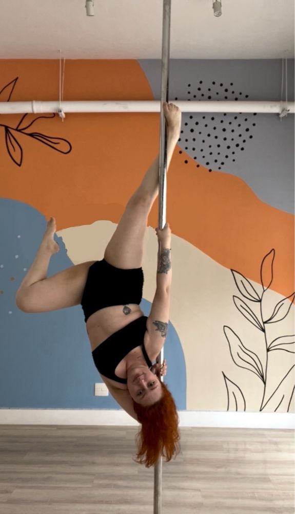 Mulher praticando pole dance. Na pose, ela está de cabeça para baixo, com o cabelo ruivo solto e esvoaçante. Veste top e shorts pretos. A pose consiste em se pendurar com o braço direito na barra, enquanto apoia o pé direito para estabilidade e solta o restante do corpo em direção ao chão, dobrando o joelho esquerdo com a ponta do pé apontada para cima. A mão esquerda passa por trás do pescoço, indo de encontro a barra e oferecendo maior estabilidade. O movimento se chama Possêidon!