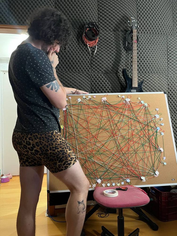 Leo, um homem branco com cabelos pretos cacheados na altura da orelha está pensativo na frente de um quadro marrom com inúmeras linhas verdes e vermelhas conectando diversos círculos de papel. Ele veste uma camiseta preta com pontinhos brancos e um short de oncinha bem justo evidenciando seu saliente traseiro.