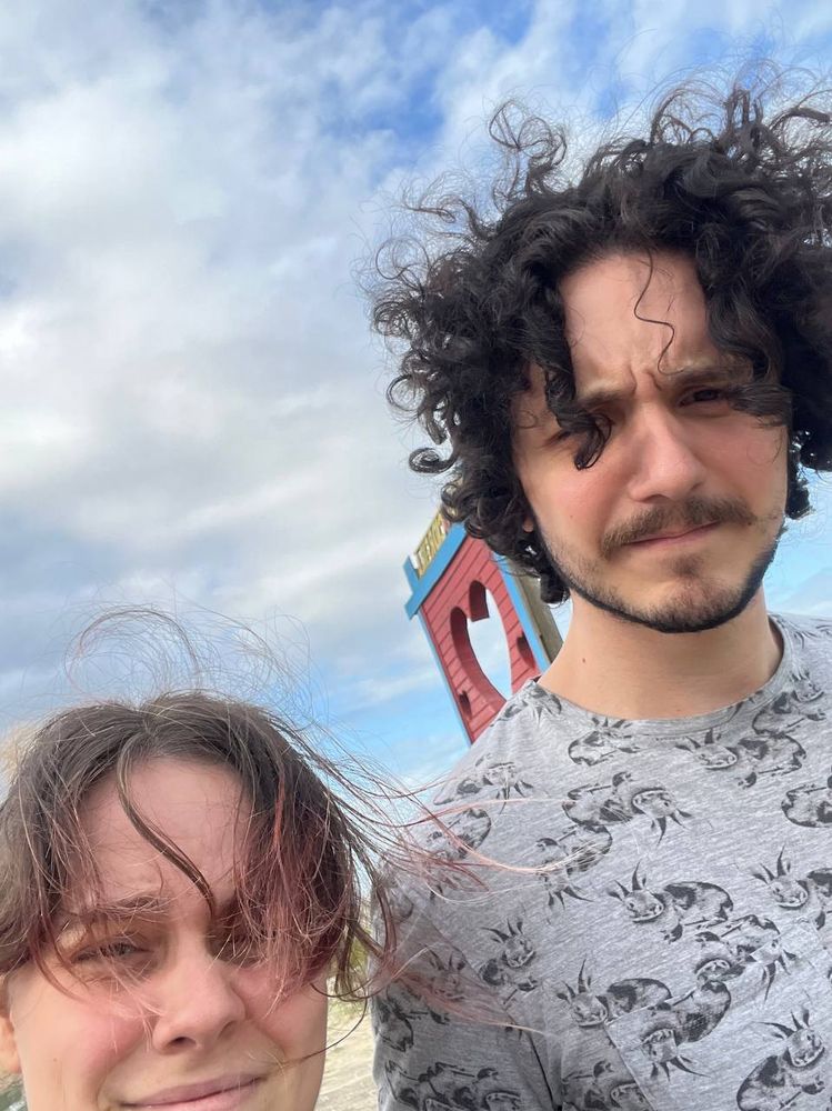 Leo, um homem branco está com os cabelos pretos e cacheados descabelados pelo vento, usa barba e uma camiseta cinza com estampa de coelhos. Vick, mulher branca também está com os cabelos castanhos de pontas rosas bagunçados do vento.