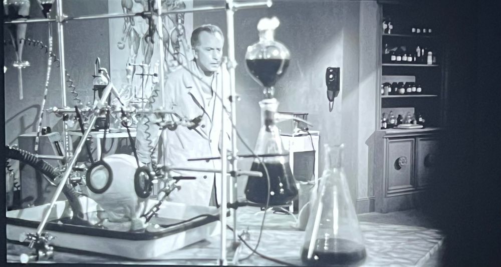 A scientist in lab stands in the background. A severed female head sets in a bloody tray in a laboratory rig, visible from behind.