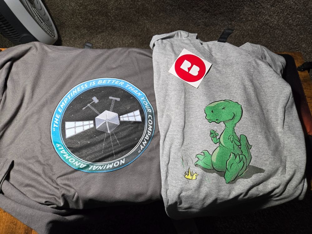 two shirts on a desk, left dark grey shirt with a space probe logo, right light grey shirt with a dinosaur logo