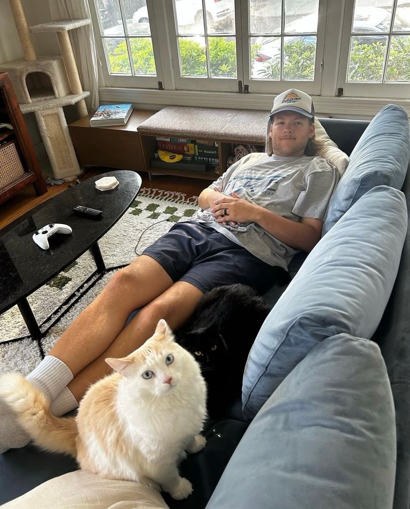 A young man lying on a blue couch with a long haired blue eyed stunning cat sitting at his feet looking at the camera