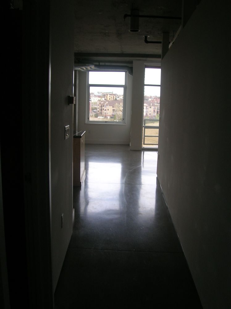 First pic taken after closing. A long dark hall with a window and a balcony at the end. Light from both shines on a grey concrete floor. The view thru the window and balcony is of a park with a green hill and beyond that other mid-rise buildings. 