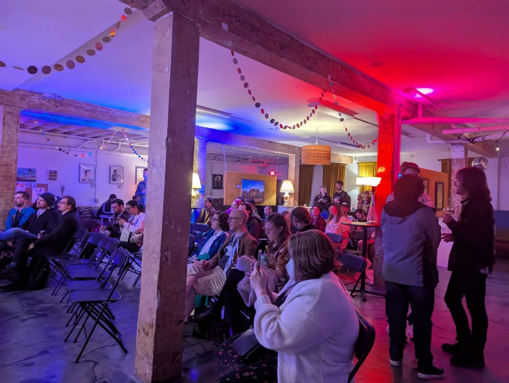 A large crowd of people sitting and listening to music in a basement with cool mood lighting. 