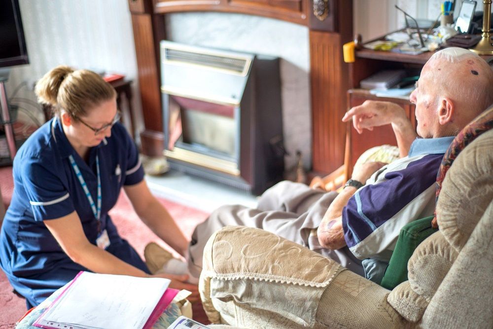 A female district nurse kneels on the floor in front of an elderly male patient in his living room. She is changing a dressing on his foot.