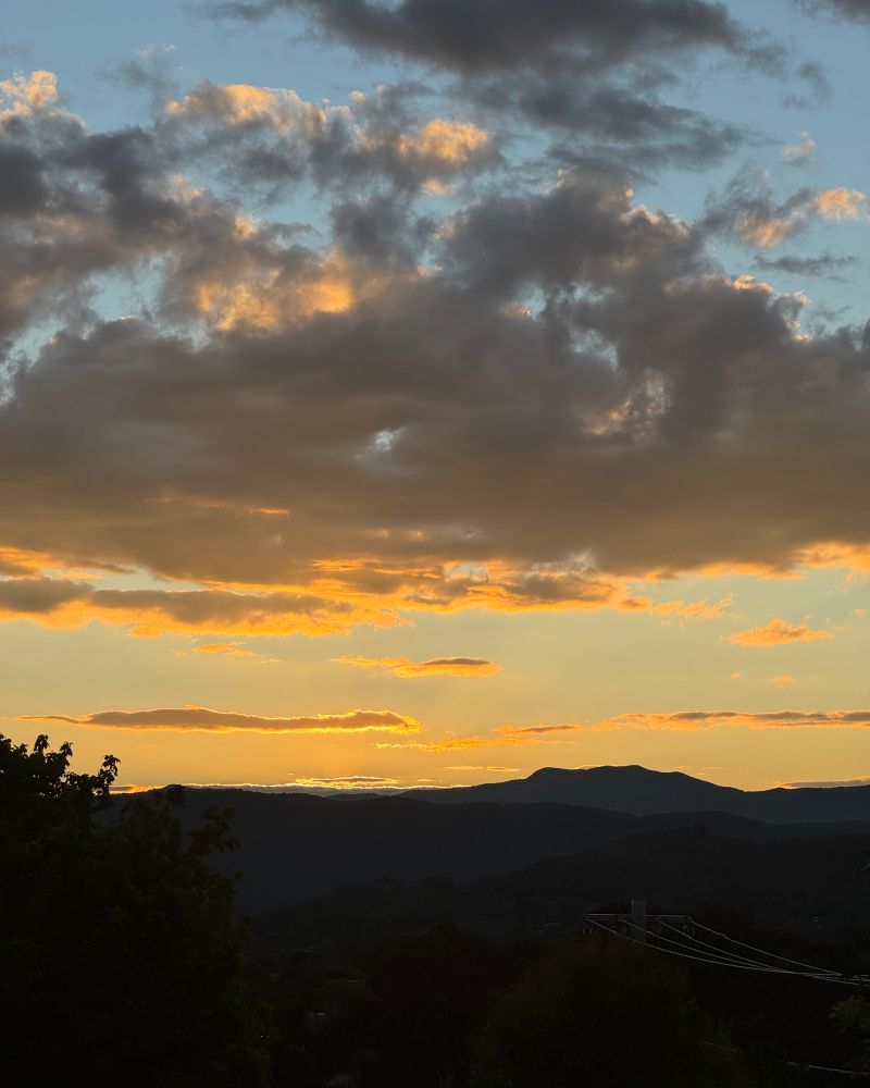 A view of Mt Tidbinbilla on sunset. Tidbinbilla is a dark bulk against a bright sky in blue, orange and grey.