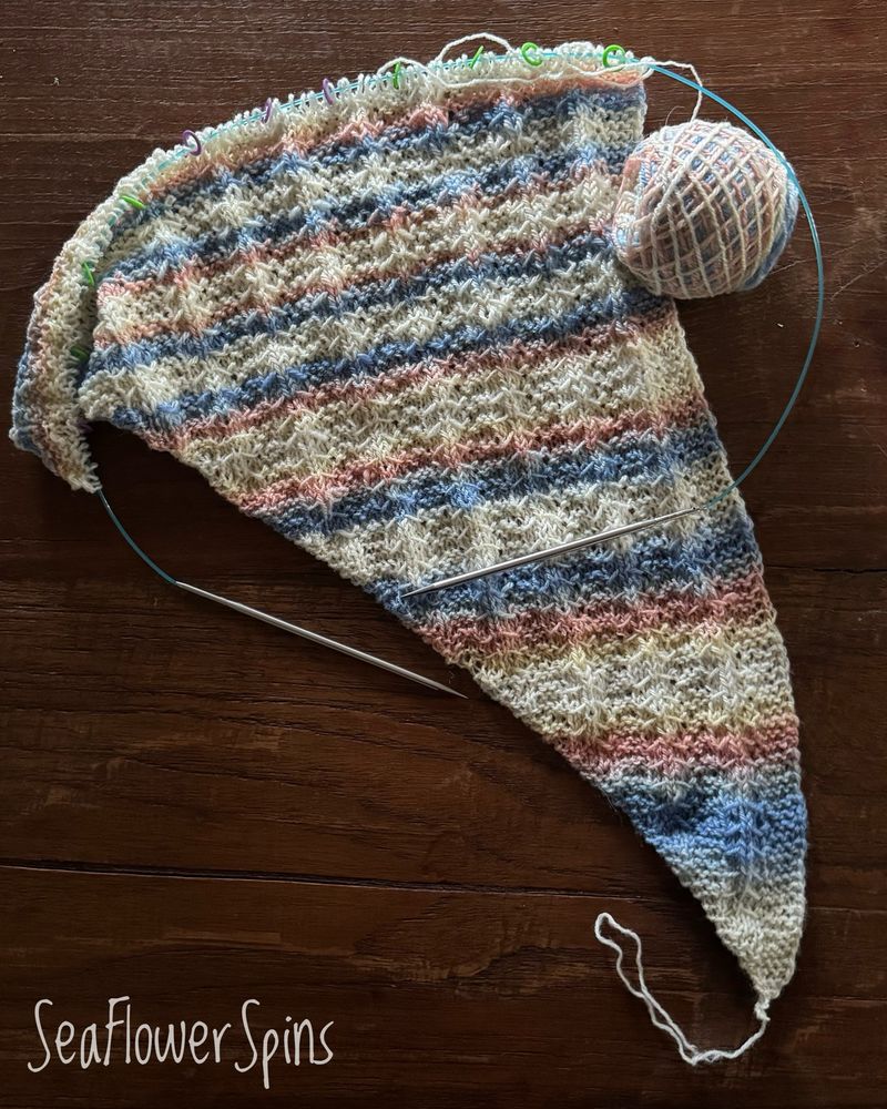 The corner of an in-progress asymmetrical shawl knit with a ball of working yarn. It lies on a dark table; the yarn is coloured white with pale blues and oranges, and a hint of yellow. 