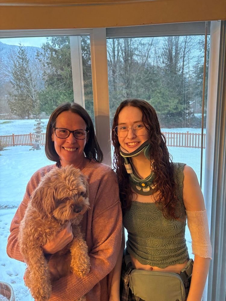 Natalie (a 19yo with reddish hair, wearing a hard collar) stands next to her mom Beth (a 50yo
With short brown hair and glasses). Beth is holding a small cavapoo and wearing a pink sweater. Natalie is wearing a green tank top and has a picc line. 