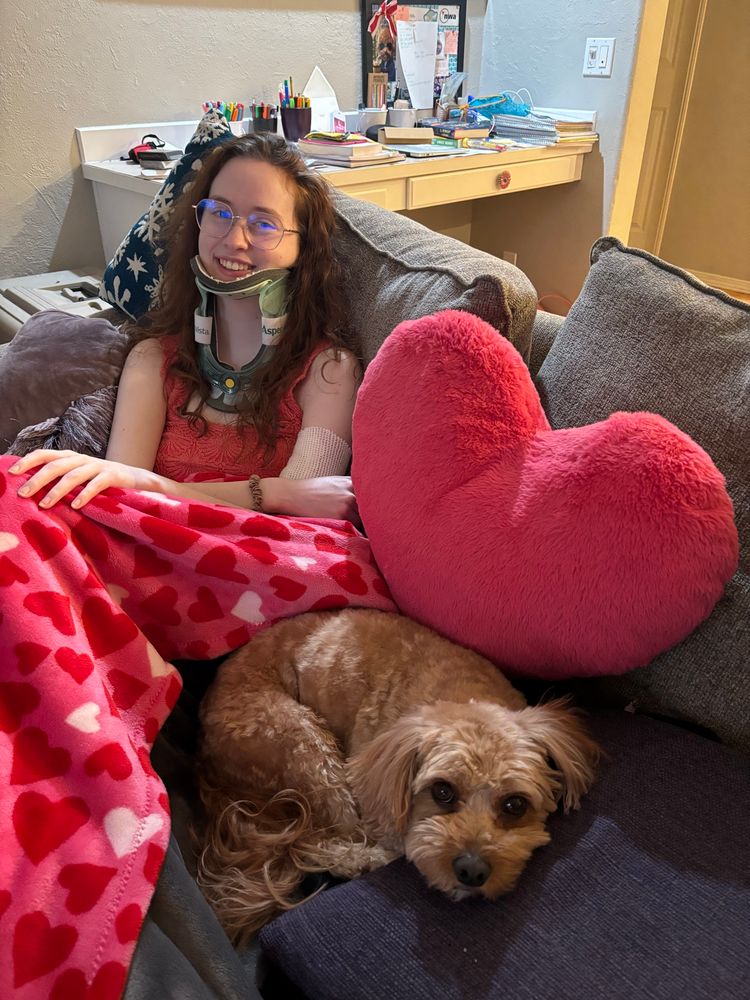 Natalie, a 19yo with reddish hair sits on a couch with a small dog next to her. Natalie is wearing a hard cervical collar. She has a blanket with hearts over her and a large heart pillow next to her. 