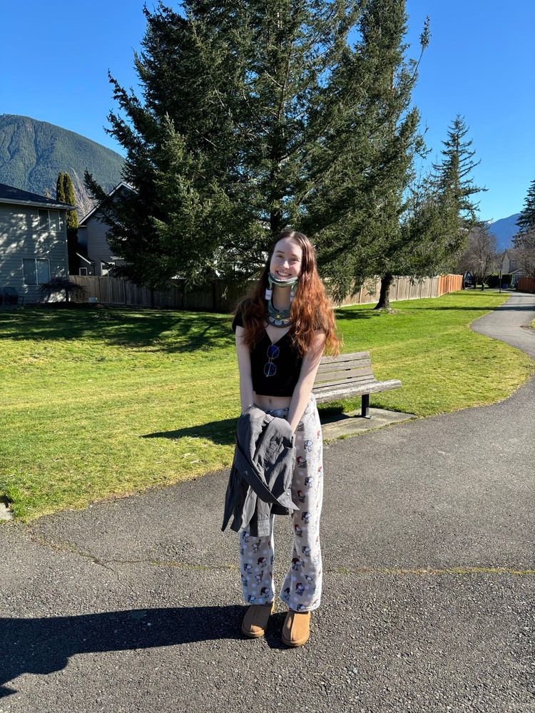 Natalie, a slender 19 yo with redish hair stands outside in the sun in pajama bottoms and a black tshirt. She is smiling and there is a tree and mountain behind her.
