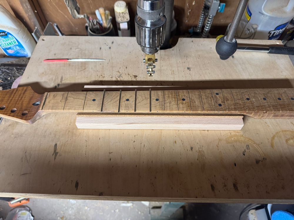 A guitar neck sitting on a drill press table. There are no frets in some slots of the neck. 