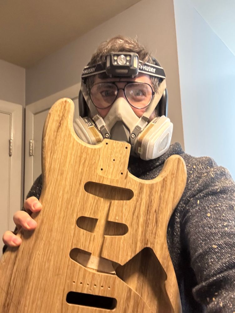 A man holding a guitar body, wearing a respirator, safety glasses and a head light. 