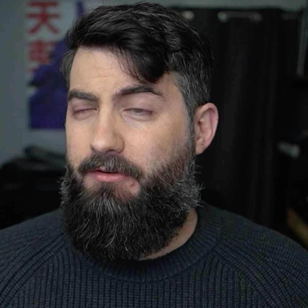 A bearded man mid speaking with a weird look on his face. A single frame from a longer video 