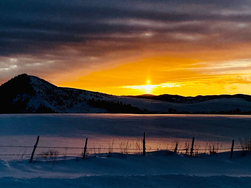 The photo captures a serene sunrise over a snow-covered landscape. The sun is low on the horizon, casting a warm golden glow across the sky and reflecting faintly on the snowy ground. Silhouetted against the light are rolling hills and a fence with posts, while dark clouds add a dramatic contrast to the vibrant hues.