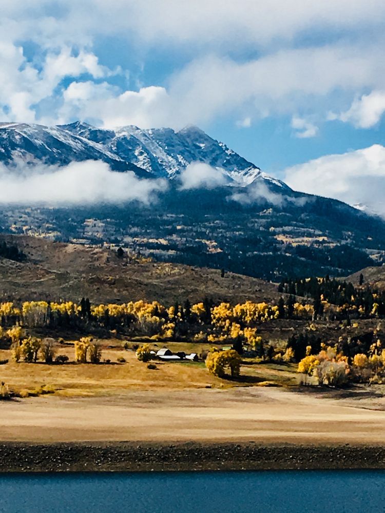 This photo captures a serene autumn mountain landscape, featuring a bright blue sky and snow-dusted peaks partially shrouded in fluffy white clouds. The mid-ground showcases a vibrant band of golden-yellow trees, interspersed with darker evergreens, framing a small cluster of buildings nestled within the valley. In the foreground, a calm body of water stretches across the bottom of the frame, reflecting the subdued light of the scene. 