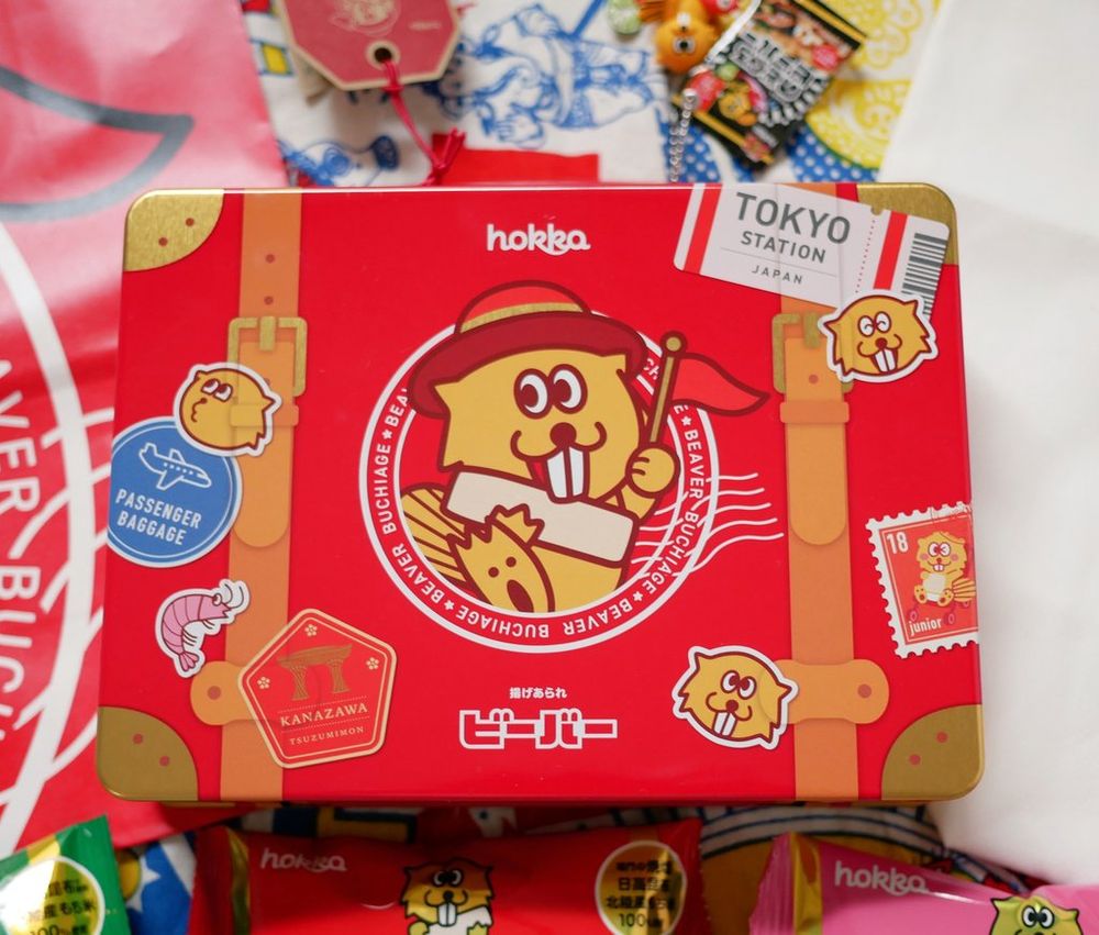 缶素材のトランクのアップ。ビーバーのキャラクターの絵が真ん中にあり、取っ手とベルトが付いた旅行鞄のようなデザインになっている。パスポートスタンプのようなシールのような柄がちりばめられている。