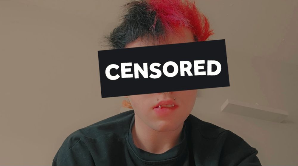 A selfie of me wearing a black shirt and biting my lip a little. I have two vertical top lip piercings called "angel fangs" and there's a big black rectangle blocking out a big portion of my face and it says "censored"
