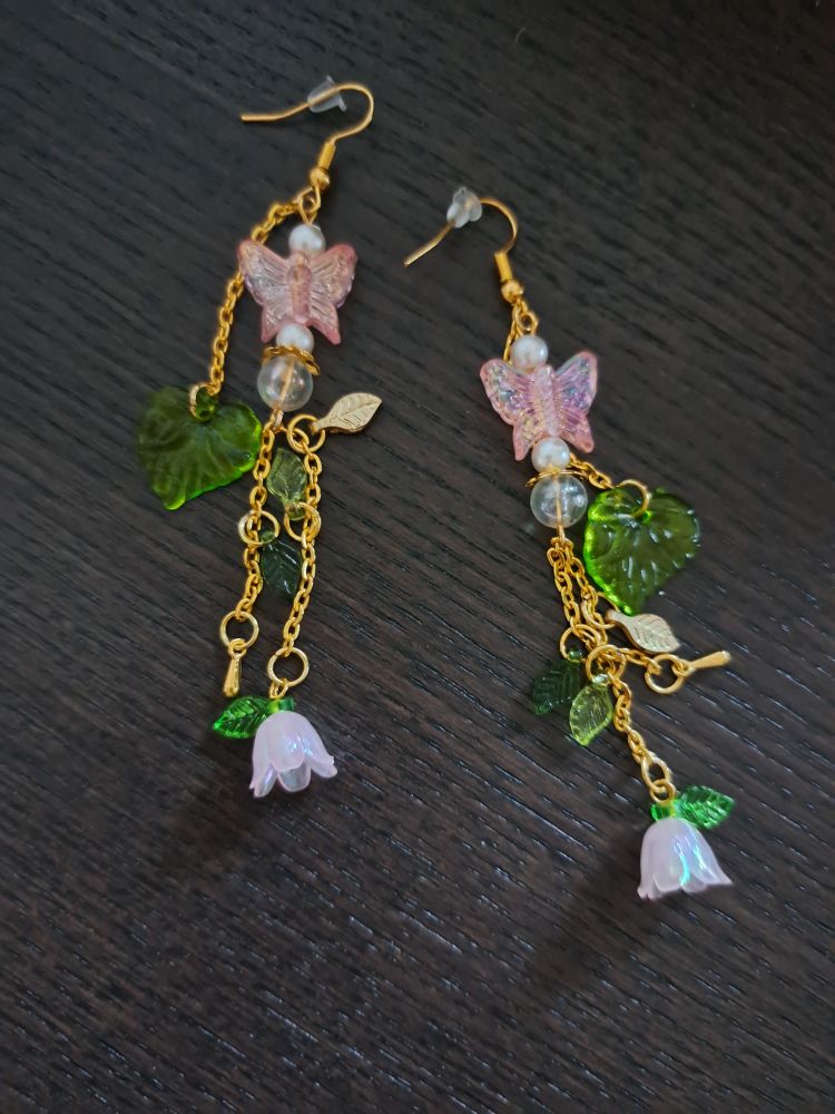 A set of long earrings. On small golden chains hang pink flowers, green leaves and there's a pink butterfly on top. It lies on top of a table. 