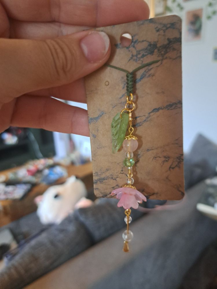 A phone charm with a pink, lotus-like flower and light pink and green pearls, as well as a leaf charm