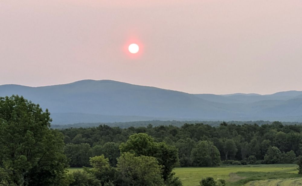 Morning sun with pink halo seen over mountains with trees and fields in foreground.