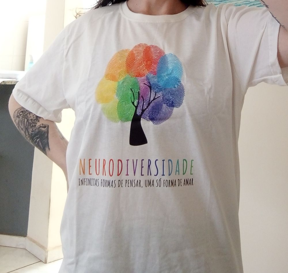 Uma pessoa vestindo uma camiseta com a frase: neurodiversidade: infinitas formas de pensar, uma só forma de amar com o desenho de uma árvore acima da frase, onde os galhos são representados com a união de representações de impressões digitais coloridas