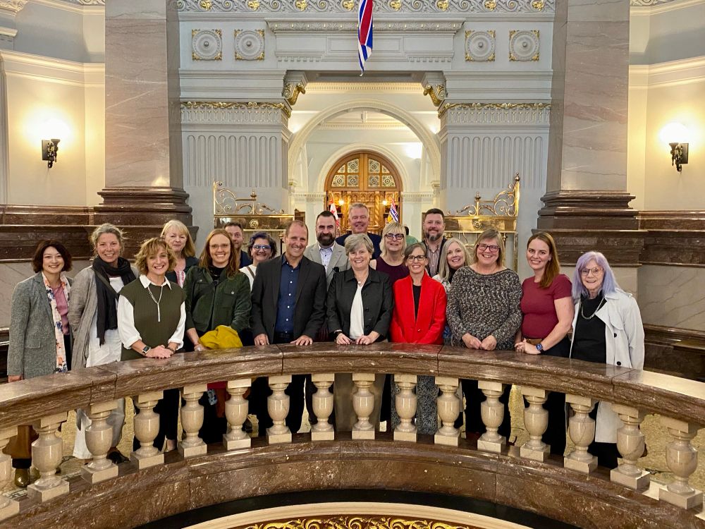 Group of ~20 physicians, support staff, and B.C. Minister of Health standing around the circular stone balustrade in the rotunda at the B.C. Legislature, facing the camera and smiling. The ornate doors to the chamber of the Legislature are visible in the background. 