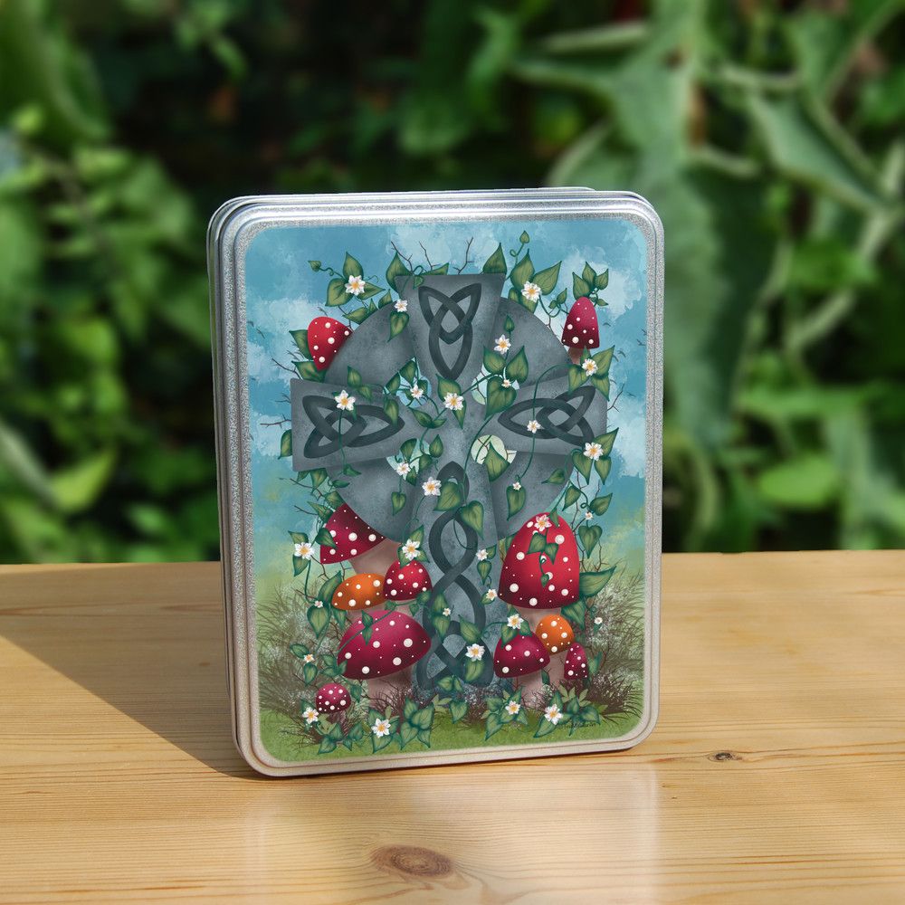 Rectangular metal tin: A grey stone Celtic cross, inlaid with Celtic knots, stands in long grass surrounded by red and orange spotted toadstools. Leaves and small white flowers twine upwards and through the cross, and more toadstools appear near the top. There's a mottled blue sky in the background.
