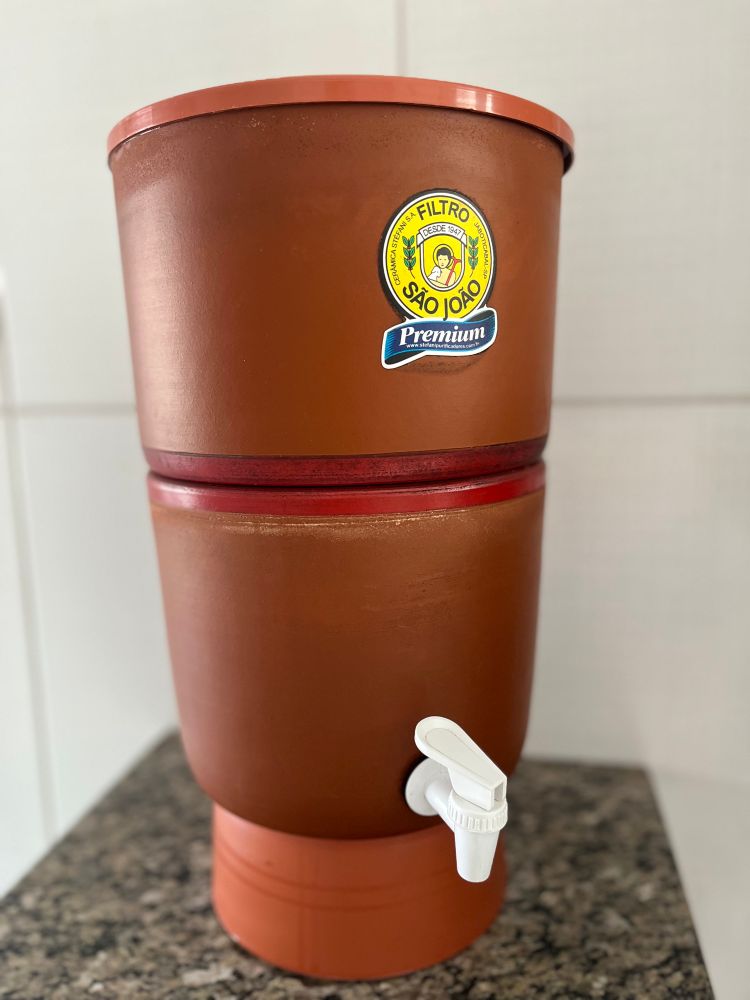 Keramische Wasserfilter in roter Keramik mit einem weißen Plastikhahn in weiß. Auf dem oberen Teil ist ein gelber Aufkleber. Die Wand ist weiß gekachelt. Der Filter steht auf einer grauen Marmorplatte 